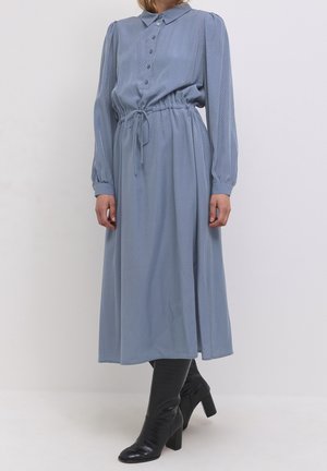 Vrouw draagt een lichtblauwe midi-jurk met lange mouwen, kleine stippen, een aantrekkoord in de taille en zwarte kniehoge laarzen met hak.