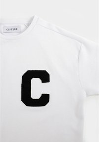 Hvit bomull T-skjorte med et stort svart filt bokstav "C" på brystet. Rund hals og korte ermer. Enkel, ren design.