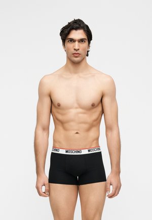 Jeune homme aux cheveux courts et foncés porte un boxer noir Moschino sur un fond uni gris clair, debout face à l'avant.