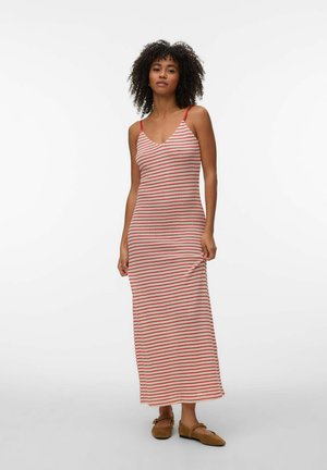 Femme aux cheveux bouclés portant une robe sans manches, longue jusqu'aux chevilles, rayée rouge et blanche, et des chaussures plates marron, debout devant un fond blanc.