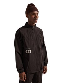 Mand iført en brun strikket hue og sort lynlåsjakke med "223" og "HALO" trykt på brystet, står mod en hvid baggrund.