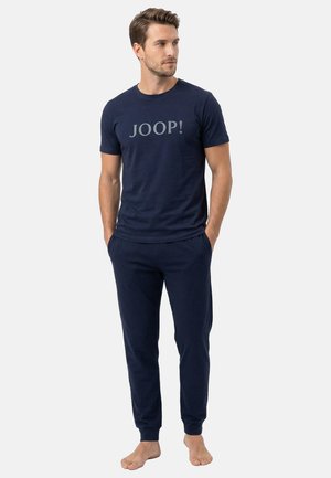 Mężczyzna w granatowej koszuli z krótkim rękawem z napisem "JOOP!" i pasujących granatowych spodniach, stojący boso z rękami w kieszeniach.