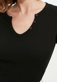 Svart ribbad stickad topp med V-ringning och tre knappar vid halslinjen. Slät yta med figurnära design.