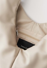 Interno della borsa beige con foderatura liscia e texture, una tasca con zip e un'etichetta in tessuto nera con testo bianco.