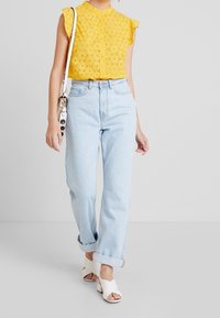 Yellow eyelet blouse with ruffled sleeves, paired with light blue high-waisted jeans. White heeled mules. Bag with decorative strap.