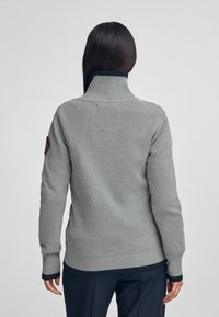 Szary sweter dzianinowy z wysoką szyją i ściągaczami. Posiada boczne rozcięcia oraz kontrastowe ciemne wykończenie. Zawiera naszywkę na lewym rękawie.