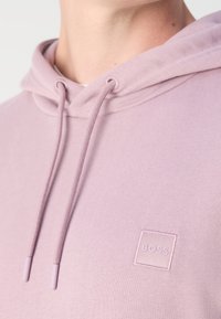 Gros plan d'une personne portant un sweat à capuche violet clair avec des cordons et un petit patch logo "BOSS" sur la poitrine.