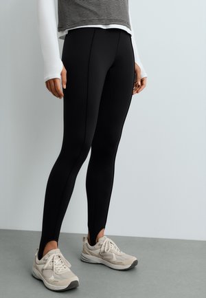 Personne portant un legging noir slim avec étriers pour les pieds et des chaussures de course blanches, debout devant un mur clair uni.