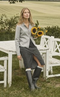 Grau zip-up Pullover, dunkler Jeansrock mit einem Schlitz vorne und schwarzen Gummistiefeln. Hält einen Strauß Sonnenblumen. Außeneinstellung.