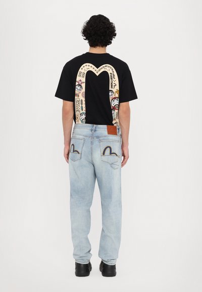 Melna T-krekls ar krāsainu grafisko dizainu, kurā ir liels arcs aizmugurē, apvienots ar gaiši ziliem džinsiem un melnu apavu.