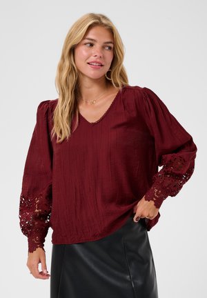 Kvinde med langt blondt hår iført en bordeauxbluse med blondeærmer og sort nederdel, der ser til siden med et let smil.