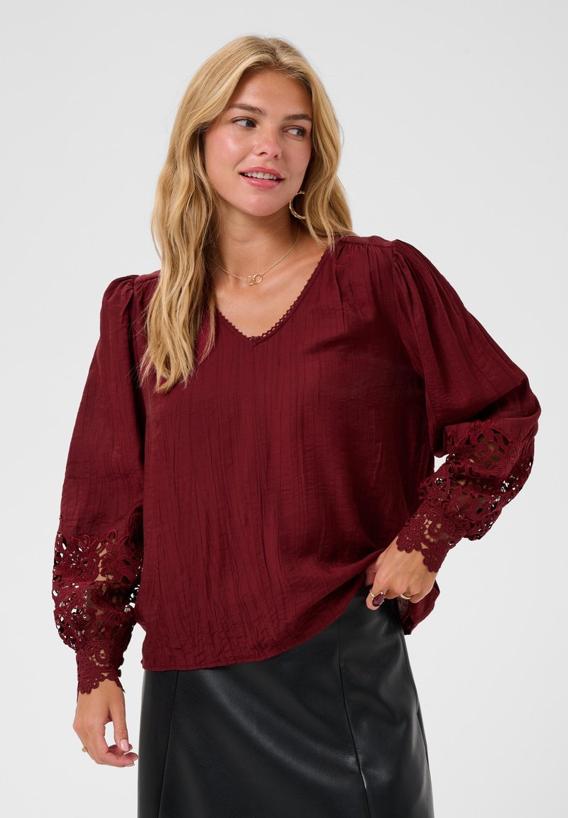 Femme aux longs cheveux blonds portant un chemisier bordeaux à manches en dentelle et une jupe noire, regardant sur le côté avec un léger sourire.