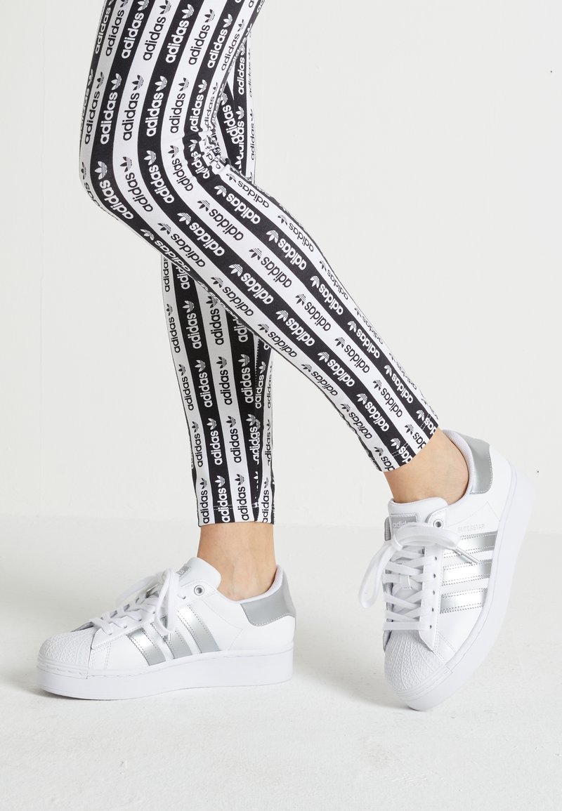 Nohy v černobílých legínách s logem Adidas a bílých teniskách Adidas Superstar se stříbrnými pruhy, na jednoduchém bílém pozadí.