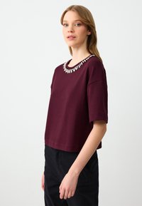 Bordeaux katoenen T-shirt met korte mouwen en een ronde halslijn, versierd met heldere kraalversieringen. Gecombineerd met zwarte broek.