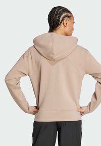 Žena s opletenými vlasmi v béžovej mikine a čiernych nohaviciach, stojaca so založenými rukami v páse, otočená chrbtom k fotoaparátu na jednoduchom pozadí.