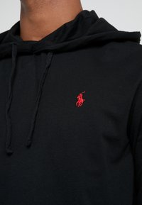 Svart huvtröja i mjukt material med dragsnöre, med en liten röd broderad logotyp på vänster bröst.