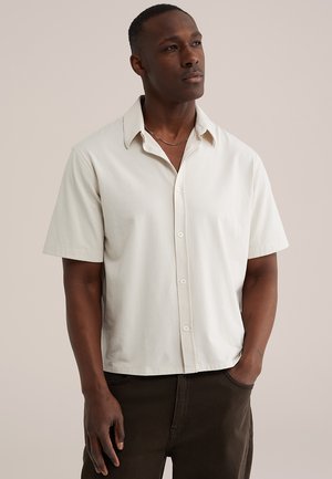 Dunkelhäutiger Mann, trägt ein kurzärmliges weißes Hemd mit Knöpfen und dunkle Hose, steht mit einer Hand in der Tasche und blickt leicht nach oben.