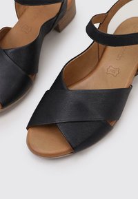 Sandalias de cuero negro con un diseño de tiras cruzadas, punta abierta y un tacón de madera bajo. El interior es de cuero marrón claro con un acabado texturizado.