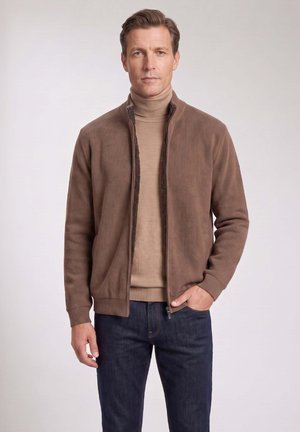 Mann trägt braune Jacke mit Reißverschluss über beigem Rollkragenpullover und dunkelblauen Jeans, steht mit einer Hand in der Tasche.