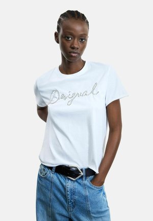 Giovane donna con capelli intrecciati che indossa una t-shirt bianca con la scritta "Desigual" ricamata e jeans blu a vita alta con cintura nera.