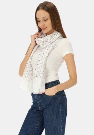 Młoda kobieta ubrana w biały krótki rękaw, niebieskie dżinsy oraz biały szal z małymi wzorami kotwic, pozująca z jedną ręką w kieszeni.