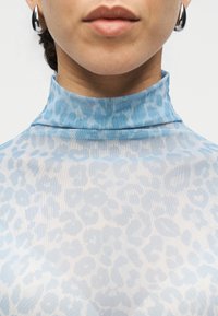 Ανοιχτό γαλάζιο, διάφανο τοπ με ψηλό λαιμό και μοτίβο λεοπάρδαλης. Διαθέτει ψηλό γιακά και ομαλή, υφασμάτινη υφή. Κυκλικά ασημένια σκουλαρίκια.