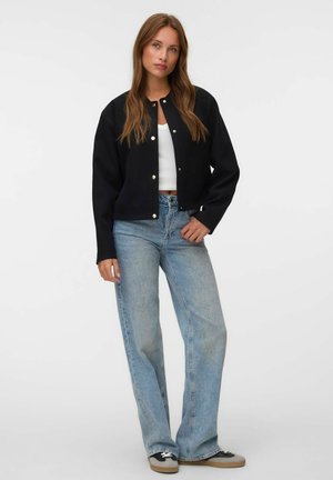 Femme portant une veste noire boutonnée, un crop top blanc, un jean bleu clair et des baskets noires et blanches, debout devant un fond uni.