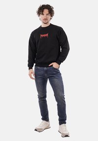 Felpa nera con testo grafico rosso sul petto, abbinata a jeans blu e sneakers bianche, in piedi contro uno sfondo semplice.