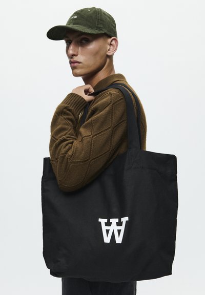 Schwarze Tragetasche mit weißem Logo, aus strapazierfähigem Stoff. Verfügt über lange schwarze Träger und eine weiche Textur. Getragen mit einem braunen Ringelmuster-Pullover.