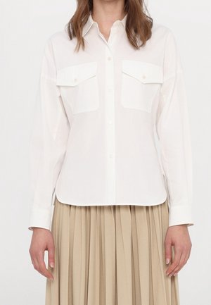 Femme portant une chemise blanche à boutons avec deux poches avant et des manches longues, associée à une jupe plissée beige.