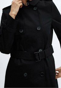 Schwarzer Trenchcoat mit doppelreihigem Design, großen Knöpfen, einem Gürtel in der Taille und einem Kragen. Hergestellt aus einem glatten, strukturierten Stoff.