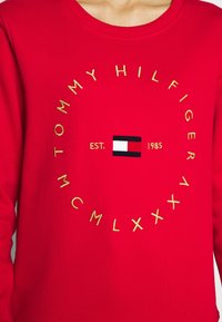 Rode sweatshirt met gele geborduurde tekst "TOMMY HILFIGER MCMLXXXV" die de Tommy Hilfiger vlag logo omringt en de tekst "EST. 1985".