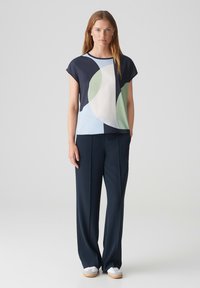 Blusa geometrica blu navy e azzurra con maniche corte, abbinata a pantaloni larghi blu navy e sneakers bianche.