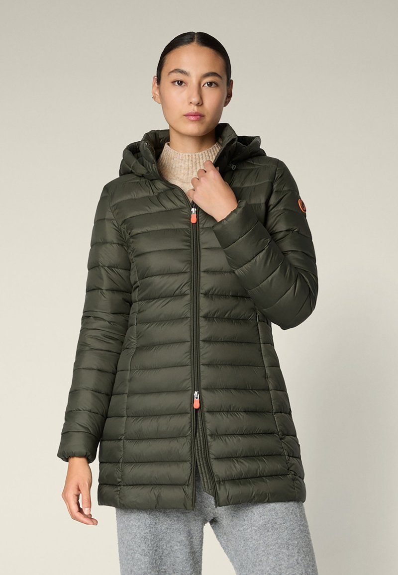 Frau trägt eine lange, olivgrüne, gesteppte Jacke mit Kapuze, die zugezippt ist, kombiniert mit einem beigefarbenen Rollkragenpullover und grauen Hosen.