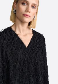 Fekete textúrájú felső V-nyakkal, egyedi, bolyhos anyagmintázattal. Ezüst fülbevalók kontrasztot adnak. Simább háttér.