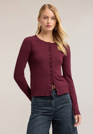 Woman wearing a fitted burgundy buttoned cardigan and dark jeans with loose blonde hair and large gold earrings against a plain background.