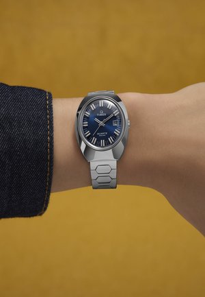 Handgelenk mit einer silbernen Timex Quarz-Uhr, die das Datum "14" und die Uhrzeit 10:10 anzeigt, vor einem senfgelben Hintergrund.