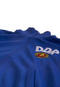 Blauwe zip-up jas met hoge kraag, 'DDR' lettering en een embleem met een goud en rood cirkelontwerp. Gladde stof structuur.