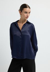 Camicia in satin blu navy con colletto, maniche lunghe e chiusura con bottoni. Texture liscia e vestibilità comoda, abbinata a pantaloni neri.