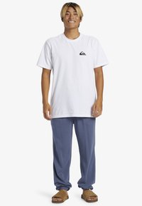 Vit bomullst-shirt med en logotyp på framsidan, parat med blå mjukisbyxor med elastiska muddar och bruna slidesandaler på en neutral bakgrund.