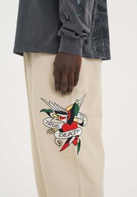 Beżowe spodnie z dużym, kolorowym haftowanym wzorem przedstawiającym ptaka i serce, z napisem "PRAWDA DO ŚMIERCI". Widać szary koszulę z długim rękawem.