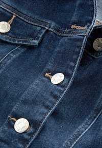 Lähikuvassa olevassa merivoimansinisessä farkkutakissa on hopeiset nepparit, näkyvät ompeleet ja haalistunut pinta, jossa on etutasku.