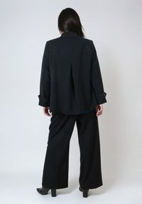 Black blazer with pleated back and rolled cuffs, paired with loose black trousers and heeled shoes, presented from the back.