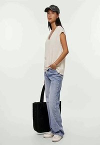 Mulher vestindo uma T-shirt bege em V, jeans azul claro, tênis brancos, boné cinza, segurando uma bolsa tote preta, de pé contra um fundo liso.