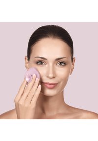 Spazzola per la pulizia del viso in silicone, lilla pallido, forma ovale con ventosa, tenuta vicino alla guancia di una donna, superficie liscia, dettagli visibili minimi.