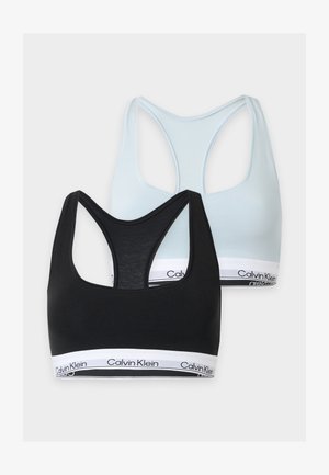 Femme debout pieds nus portant un soutien-gorge de sport Calvin Klein bleu clair et un sous-vêtement taille haute assorti sur un fond uni.
