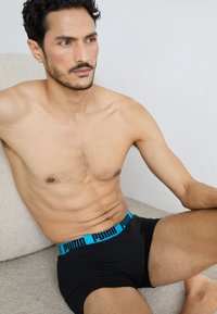 Schwarze Boxershorts mit einem blauen elastischen Bund, der den Schriftzug "PUMA" zeigt. Modell sitzt, hat kurze schwarze Haare, helle Haut und einen definierten Körperbau.