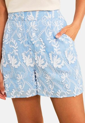 Hellblaue Shorts mit weißer Blumenstickerei, getragen mit einer Hand in der Tasche, zeigen mittlere Oberschenkellänge und strukturierten Stoff.