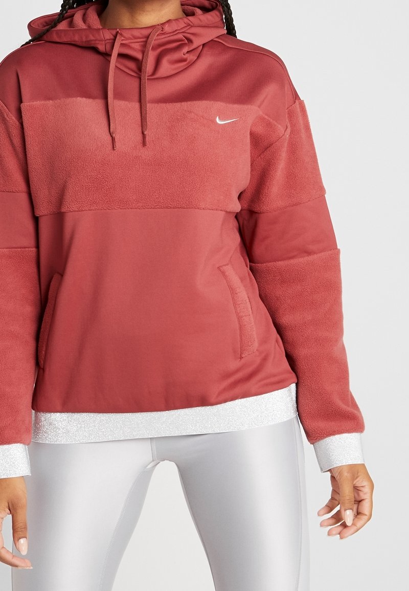 Femme portant un sweat à capuche Nike en polaire rouge avec un panneau en tissu et des poches avant, associé à un legging blanc, sur un fond uni.