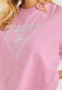 Nahaufnahme einer Person in einem pinkfarbenen Hemd mit einem großen, reflektierenden Guess-Logo, das in einem Dreieck auf der Brust gedruckt ist.